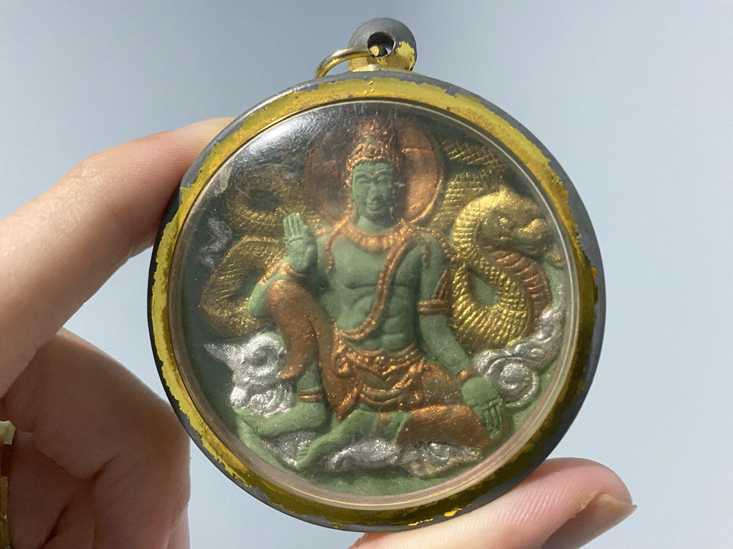 An ornate amulet depicting a Buddha, adorned with gold and encased in a glass case, with gold edges.